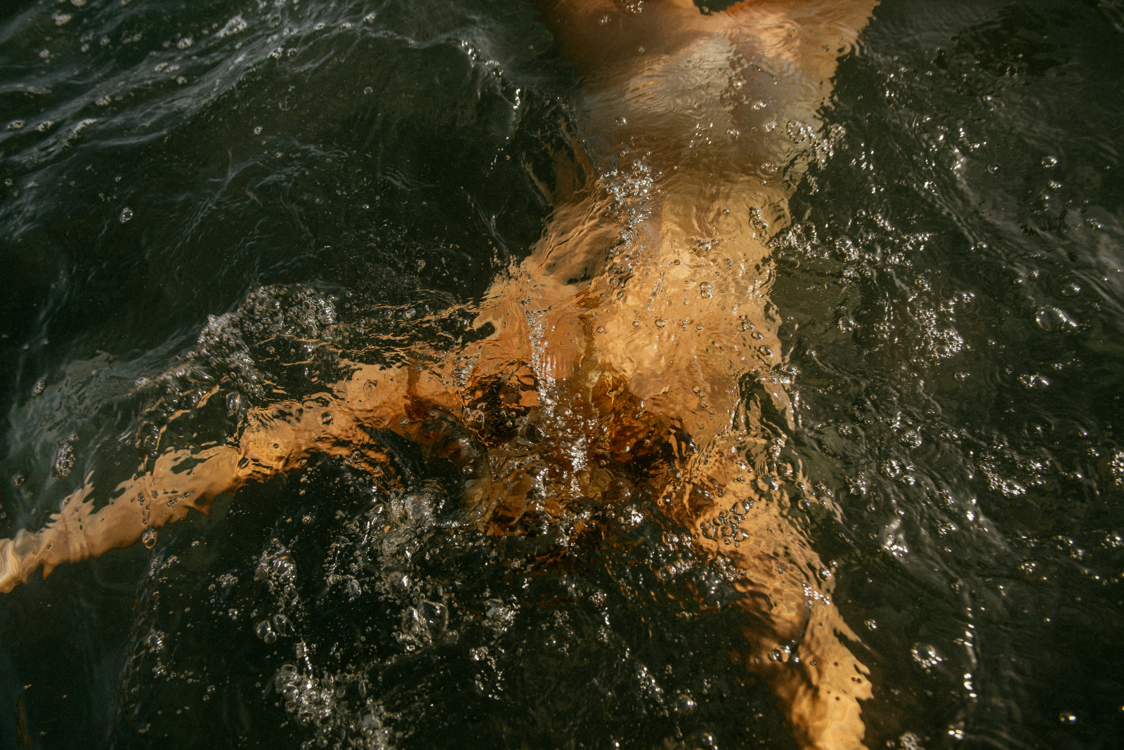 A swimmer moving through dark water at golden hour, light refracting on skin