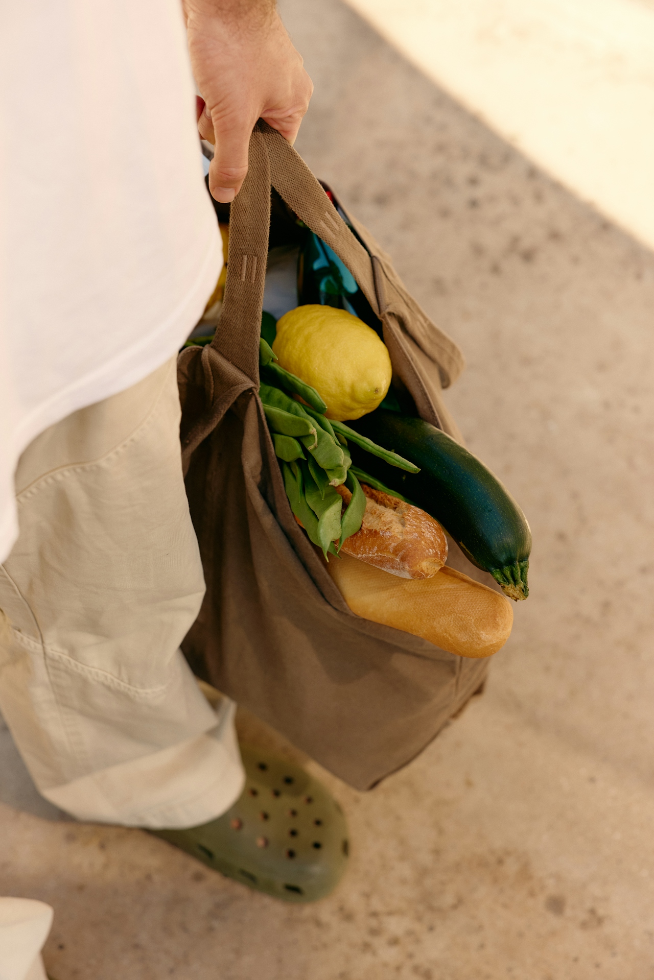 A person carrying a canvas tote bag full of fresh market produce — lemon, zucchini, green beans, and bread